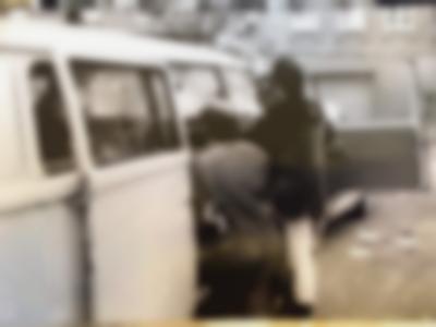 A group of three masculine-presenting people stand by the open door of a small Volkswagen bus, facing the inside.