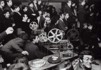 Two people sit behind a projection booth, seemingly controlling the projector. In the background, people stand up on different levels, looking in the direction the projector is facing.