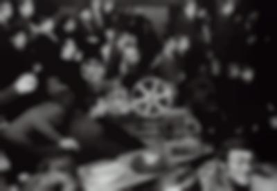 Two people sit behind a projection booth, seemingly controlling the projector. In the background, people stand up on different levels, looking in the direction the projector is facing.