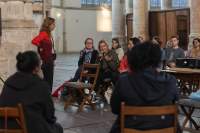 Two people sit on wooden chairs, facing an audience. Another person, stands next to a member of the audience, who is holding a microphone and gesturing with their hand.