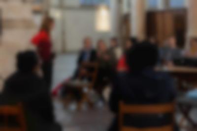 Two people sit on wooden chairs, facing an audience. Another person, stands next to a member of the audience, who is holding a microphone and gesturing with their hand.