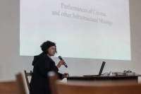 A person sits at a table, with an open laptop. They are holding a microphone to their mouth. Behind them, a large slide is being projected on the wall. The slide reads 