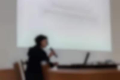 A person sits at a table, with an open laptop. They are holding a microphone to their mouth. Behind them, a large slide is being projected on the wall. The slide reads 