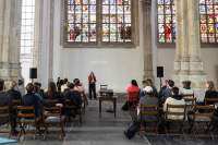 Inside a church with stained glass windows, a person stands in one of the aisles, with some text being projected behind them. Three rows of chairs, broken into two sections, are nearly full. The people look towards the presenter. In an aisle in between the chairs, there is a table with a projector, facing the presenter.