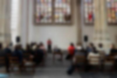 Inside a church with stained glass windows, a person stands in one of the aisles, with some text being projected behind them. Three rows of chairs, broken into two sections, are nearly full. The people look towards the presenter. In an aisle in between the chairs, there is a table with a projector, facing the presenter.