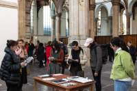 In a church, several people stand around a wooden table, with books on top. They are either holding books and paper materials, or looking at them. Behind them, a group of people is spread around the church.