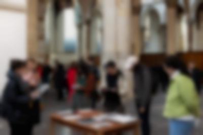 In a church, several people stand around a wooden table, with books on top. They are either holding books and paper materials, or looking at them. Behind them, a group of people is spread around the church.