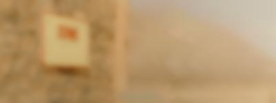 A worn-out sign, with the word Zone still visible, hangs on a building. In the background, there is a hill in an arid area. A subtitle reads: 