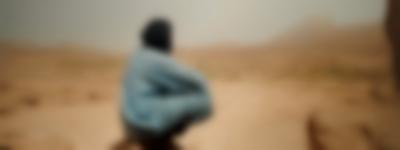 A person, squatting down, faces an arid area, with hills at the distance. They appears to be wearing a light blue long garment and a turban.