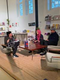 Three people sit around a low table having a discussion. Two of them hold microphones. There is a book propped up on the table.