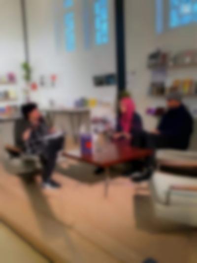 Three people sit around a low table having a discussion. Two of them hold microphones. There is a book propped up on the table.