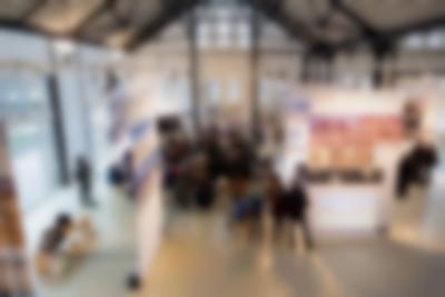 In a lofty exhibition space, a group of people gather facing a stage. Many are sitting, others stand at the back. Around them, there are large screens, with photographs printed on them. On one of the screens, a video is being projected. On stage, four people sit on chairs.