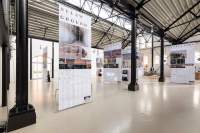 Lofty exhibition space with several large screens hanging from the ceiling. The screens have both text and images. The legible titles read: 