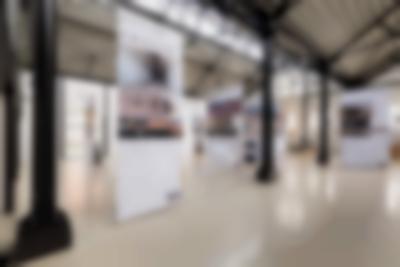 Lofty exhibition space with several large screens hanging from the ceiling. The screens have both text and images. The legible titles read: 