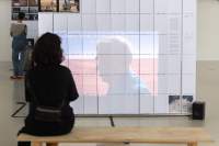 In an exhibition space, someone is sat down watching a video being projected on a large screen.