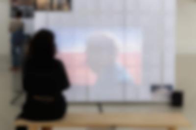 In an exhibition space, someone is sat down watching a video being projected on a large screen.