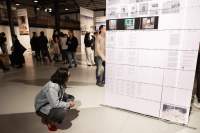Lofty exhibition space with several floor-length screens hanging from the ceiling. The screens have both text and images. It is night time and the space is filled with visitors. At the front, someone crouches down as they look at the text closest to the floor.