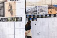 Close-up of two large screens. There are photos and documents printed on them. A person studies one of them carefully, with their arms behind their back.
