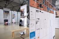 Lofty exhibition space with several large screens hanging from the ceiling. One of the screens has both images and documents with redacted information. Next to two of the other screens, there are vertical monitors playing videos. A video is being projected into another of the screens. The legible titles read: 