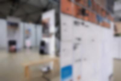 Lofty exhibition space with several large screens hanging from the ceiling. One of the screens has both images and documents with redacted information. Next to two of the other screens, there are vertical monitors playing videos. A video is being projected into another of the screens. The legible titles read: 