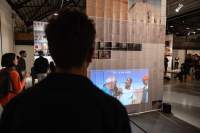 Lofty exhibition space with several large screens hanging from the ceiling. The screens have both text and images. It is night time and the space is filled with visitors. Two people watch a video being projected on one of the screens.