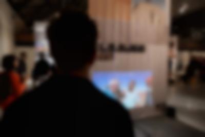 Lofty exhibition space with several large screens hanging from the ceiling. The screens have both text and images. It is night time and the space is filled with visitors. Two people watch a video being projected on one of the screens.