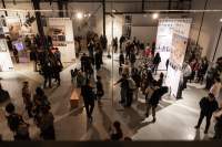 Lofty exhibition space with several large screens hanging from the ceiling. The screens have both text and images. It is night time and the space is filled with visitors.