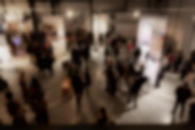 Lofty exhibition space with several large screens hanging from the ceiling. The screens have both text and images. It is night time and the space is filled with visitors.