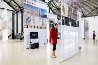 Lofty exhibition space with several large screens hanging from the ceiling. The screens have both text and images and either a video projection or a monitor. Several people occupy the space, looking at the installations.