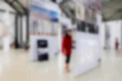 Lofty exhibition space with several large screens hanging from the ceiling. The screens have both text and images and either a video projection or a monitor. Several people occupy the space, looking at the installations.