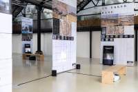 Lofty exhibition space with several large screens hanging from the ceiling. The screens have both text and images. Next to two of them, there are vertical monitors playing videos. A video is being projected into another of the screens. The legible titles read: 