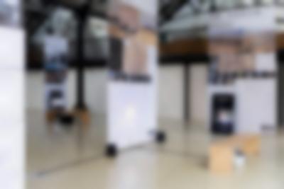 Lofty exhibition space with several large screens hanging from the ceiling. The screens have both text and images. Next to two of them, there are vertical monitors playing videos. A video is being projected into another of the screens. The legible titles read: 