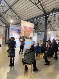 Lofty exhibition space with several large screens hanging from the ceiling. A person stands in front of one of the screens, pointing to the left. A group of people sitting in front, look in the direction the person is pointing at.
