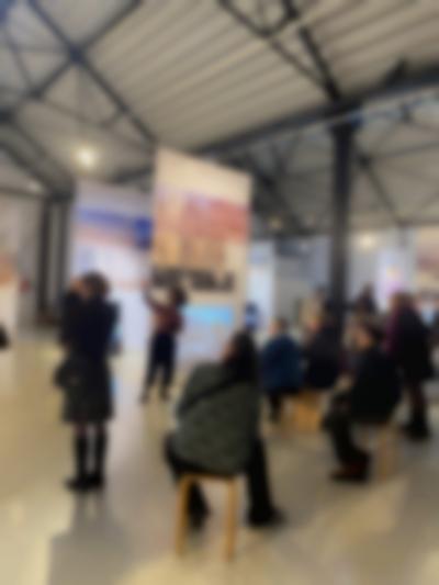 Lofty exhibition space with several large screens hanging from the ceiling. A person stands in front of one of the screens, pointing to the left. A group of people sitting in front, look in the direction the person is pointing at.