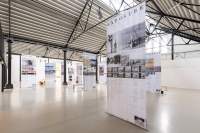 Lofty exhibition space with several large screens hanging from the ceiling. The screens have both text and images. The legible titles read: 