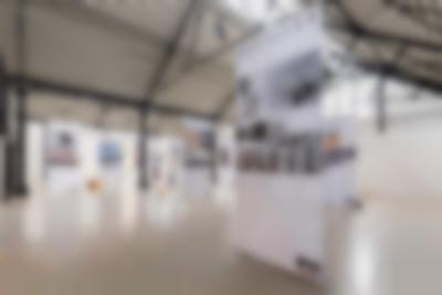 Lofty exhibition space with several large screens hanging from the ceiling. The screens have both text and images. The legible titles read: 