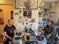 Two people are sitting across from each other at a table. They both have microphones in front of them. Behind them there is a bookshelf and a wall with posters, postcards and other decor.