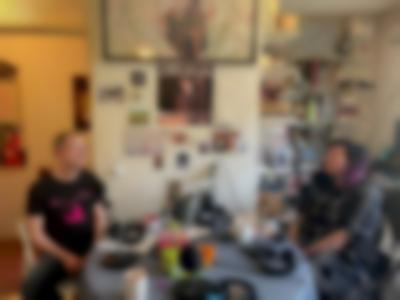 Two people are sitting across from each other at a table. They both have microphones in front of them. Behind them there is a bookshelf and a wall with posters, postcards and other decor.