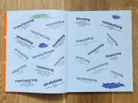 A book lies open. Across the two pages, several words in different positions and rotations have been written in black. Extra text, in a smaller font, appears underneath each word. These are words like: 