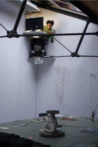A person sits in front of a suspended desk with a laptop computer, which is close to the ceiling of an indoor space. Below, there are several objects laid on a table.