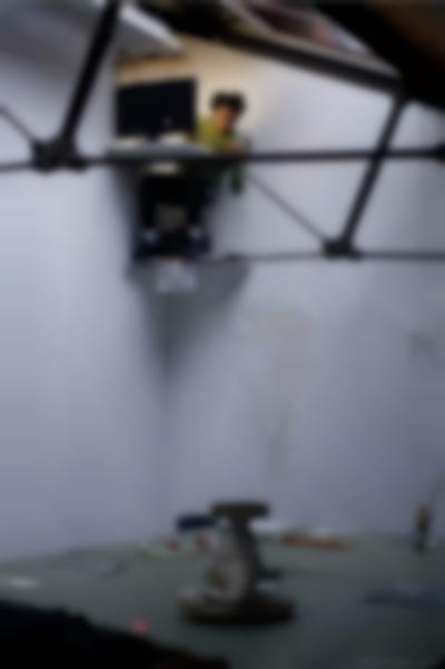 A person sits in front of a suspended desk with a laptop computer, which is close to the ceiling of an indoor space. Below, there are several objects laid on a table.