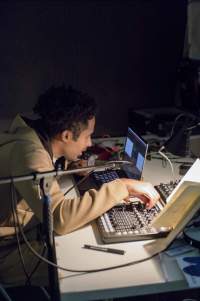 A person sits at a table, staring at a laptop and adjusting the knobs on a sound deck.