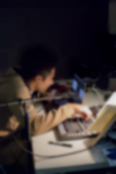 A person sits at a table, staring at a laptop and adjusting the knobs on a sound deck.