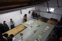 Seen from a high angle, a large table occupies most of the room. A group of people stand around the table, observing the objects that have been laid on top.