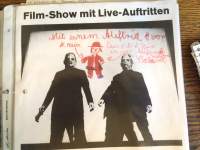 Section of a poster. It shows a black and while photograph with two people dressed in black, standing against a white background. On top of the photo, in red, there is a drawing of a clown and some handwritten notes. The event name appears above the photo.