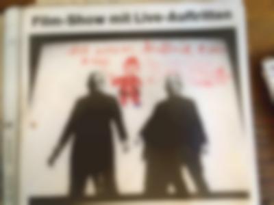 Section of a poster. It shows a black and while photograph with two people dressed in black, standing against a white background. On top of the photo, in red, there is a drawing of a clown and some handwritten notes. The event name appears above the photo.