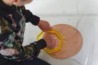 A coil spiral has been stuck to the white floor with a transparent plastic. There are two cables attached to it, one to the centre and the other to the edge. A person can partially be seen sitting next to it. With their left hand they are holding a small yellow heptagon structure against the spiral. With their right hand, they are touching the spiral through the middle of the structure.