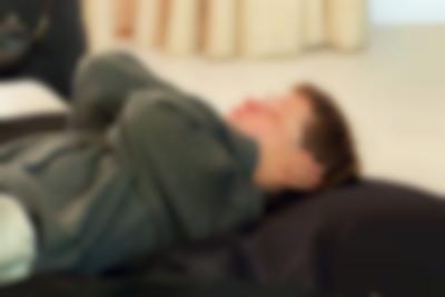 A person is lying supine on a large black cushion. They have short hair and are wearing a grey sweater. Their hands are covering their ears and their eyes are closed.