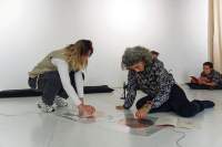 Two people seem to be playing with a sound installation consisting of two coil spirals stuck to the floor. There are cables attached to both spirals. A booklet is open next to one of them. In the background, a person is lying on the floor. They have their elbows on a pillow and are reading a book or a magazine.