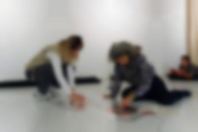 Two people seem to be playing with a sound installation consisting of two coil spirals stuck to the floor. There are cables attached to both spirals. A booklet is open next to one of them. In the background, a person is lying on the floor. They have their elbows on a pillow and are reading a book or a magazine.